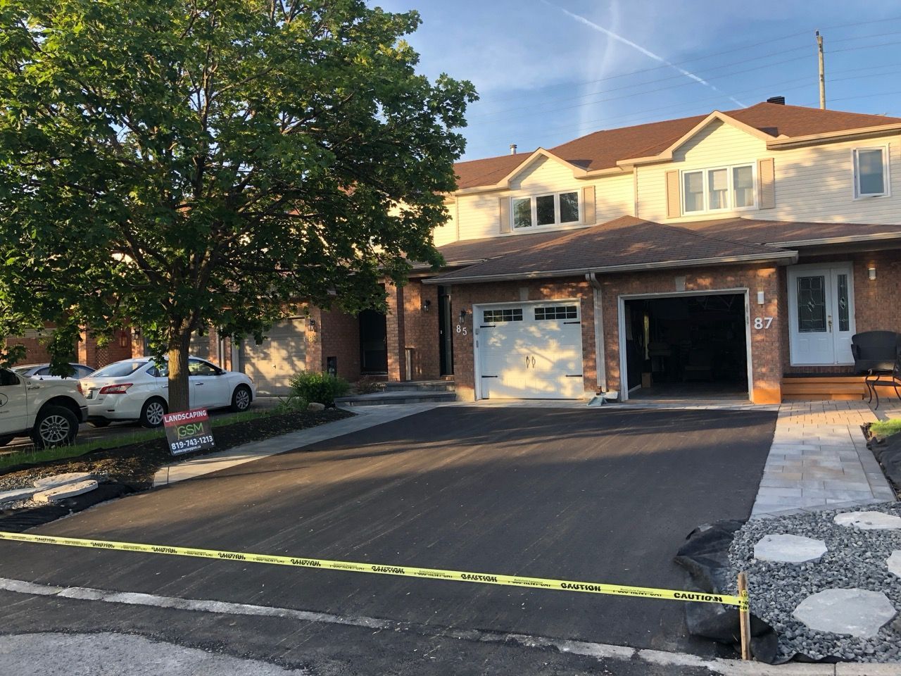 Allée asphaltée menant à une maison à deux étages avec garage ouvert. Voitures garées à gauche. Ciel visible.