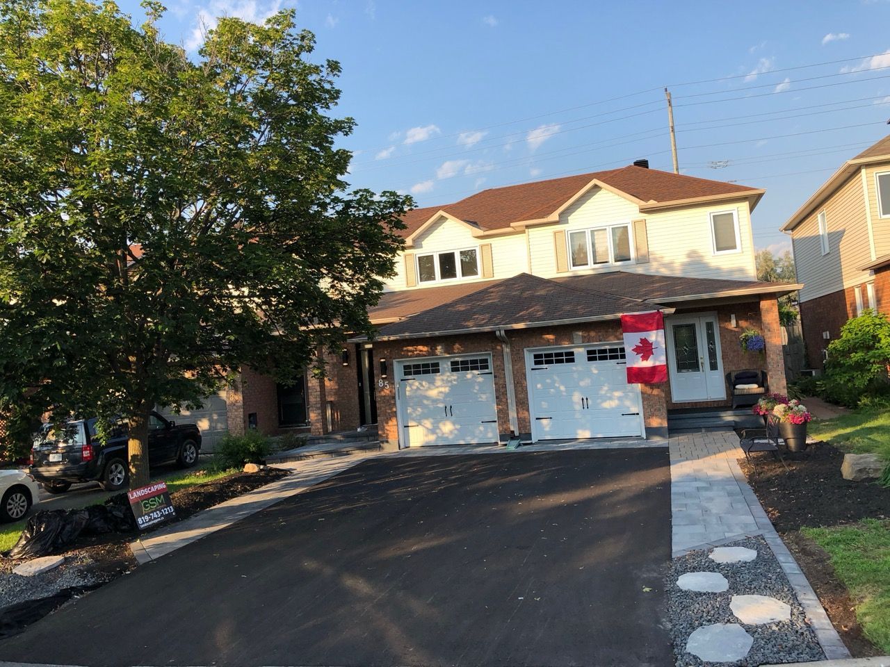 Maison avec garage double, drapeau canadien et allée récemment pavée. Arbres et ciel bleu visibles.