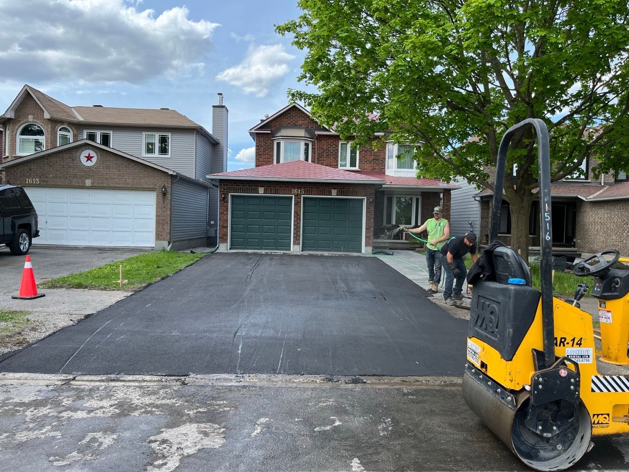 Allée asphaltée nouvellement pavée devant une maison au toit rouge avec des ouvriers et un rouleau compresseur.