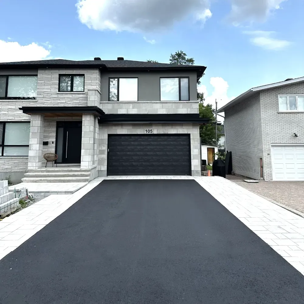 Maison moderne à deux étages avec façade en pierre grise, porte de garage noire et allée pavée.
