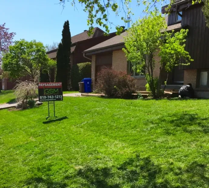Pelouse verte devant une maison en briques. On aperçoit un panneau « À vendre » et une poubelle bleue.