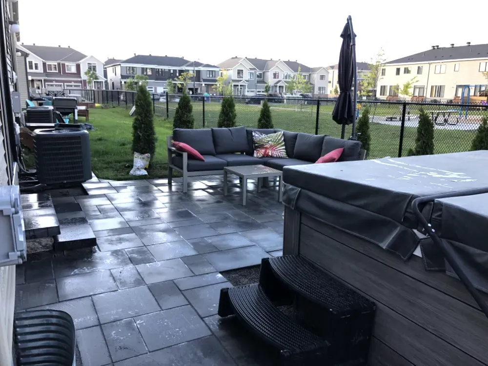 Terrasse arrière avec pavés gris, jacuzzi et salon de jardin. Pelouse verte avec maisons en arrière-plan.