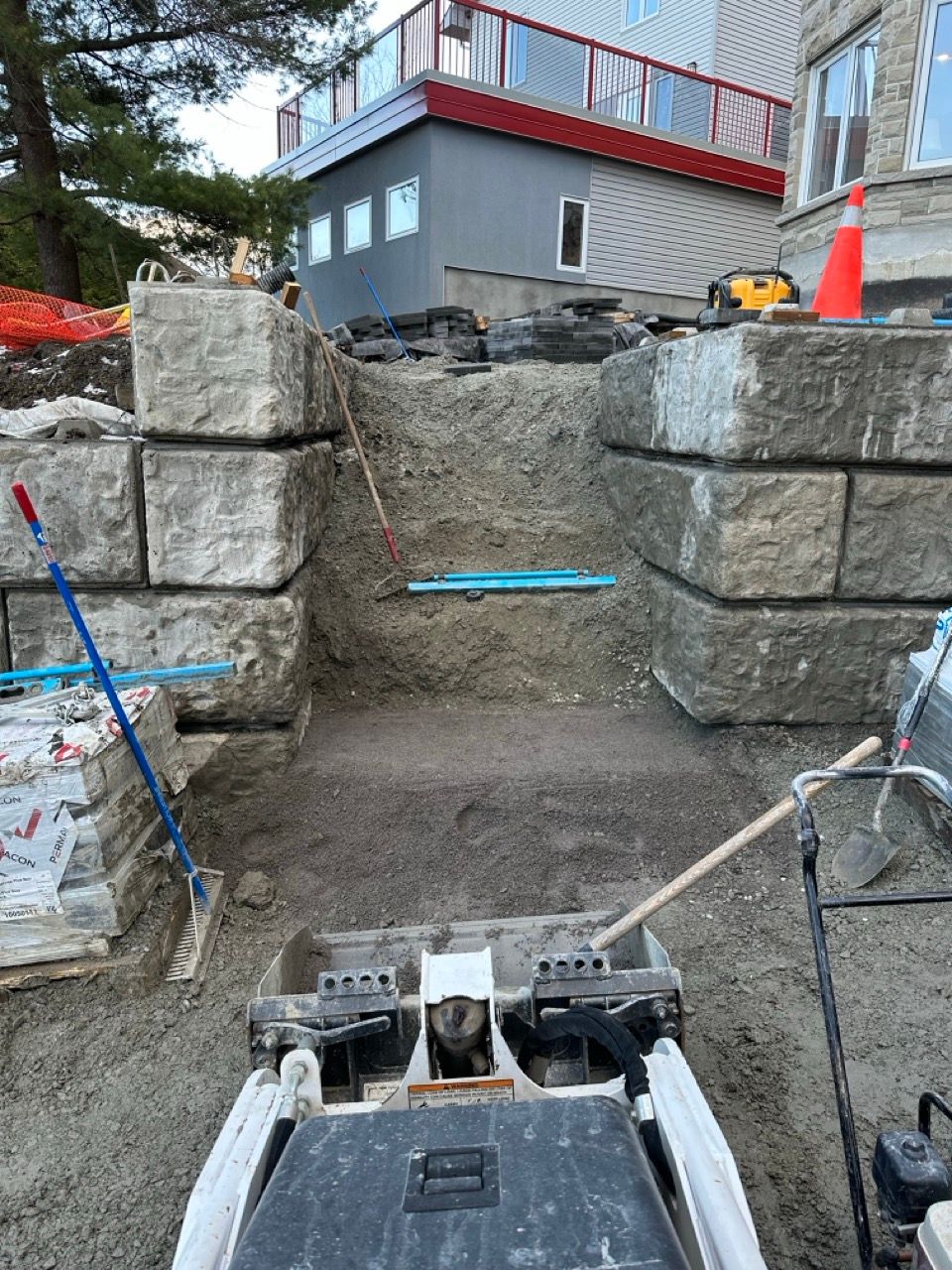 Chantier : un mur de soutènement est en construction. Une compacteuse est visible devant, entourée de pierres et d'équipements.