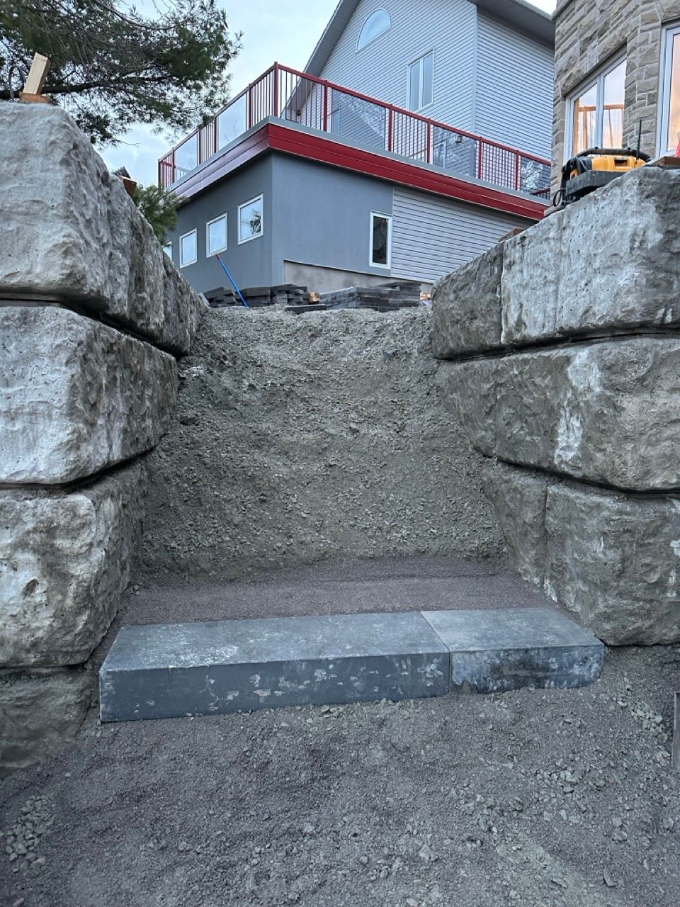 Des marches en pierre en construction, flanquées de gros blocs de pierre, mènent vers une maison grise avec un balcon rouge.