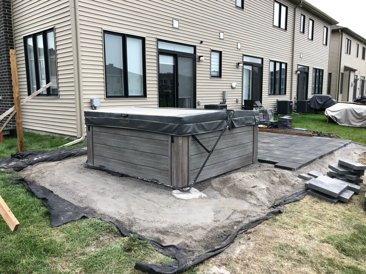Spa sur une base de sable, à côté d'un patio en construction contre une maison beige.