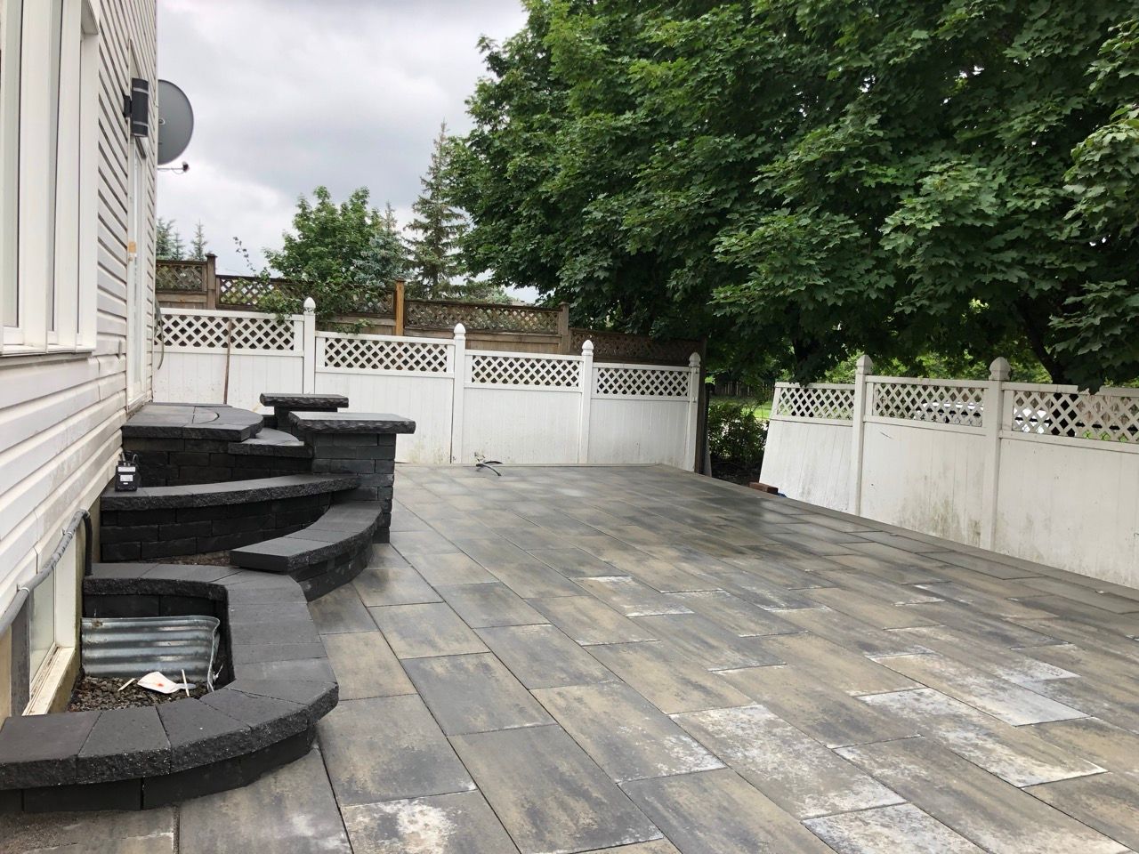 Un patio en pavés gris avec des marches, une clôture blanche et de grands arbres.