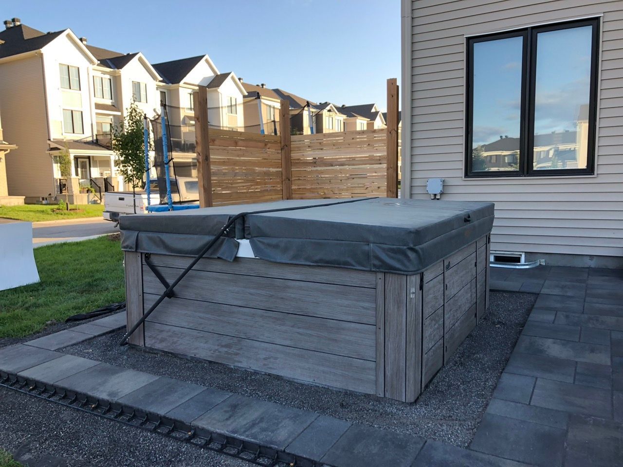 Spa avec couvercle sur un patio à côté d'une maison, avec une clôture en bois et un quartier en arrière-plan.
