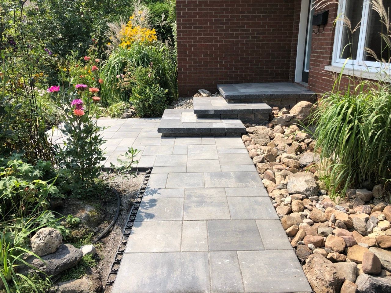 Un chemin de pavés en pierre mène à une entrée de maison avec des marches, bordée de plantes et de rochers.