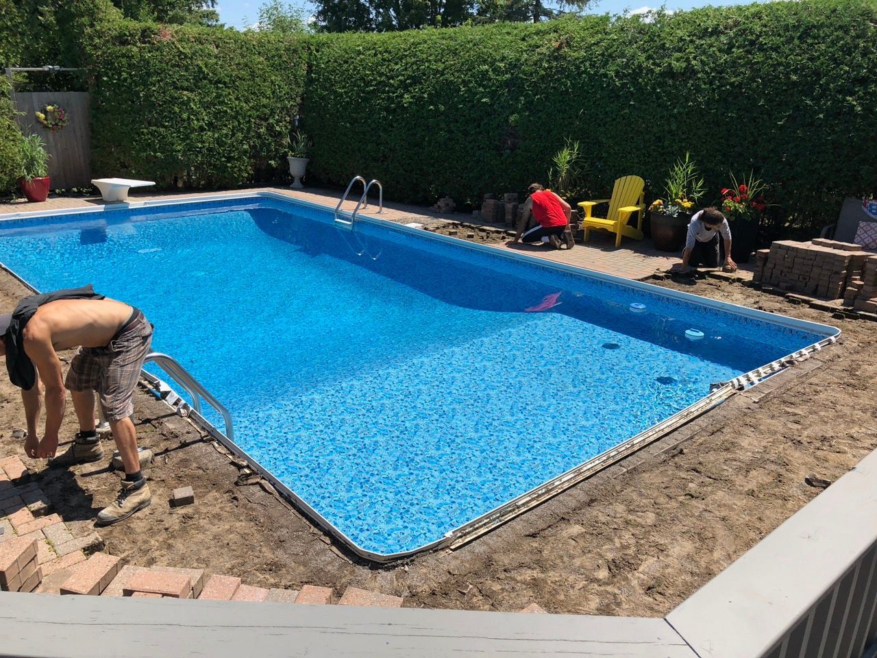 Des gens travaillent sur une piscine ; la piscine est bleue, entourée de briques brunes et de verdure.