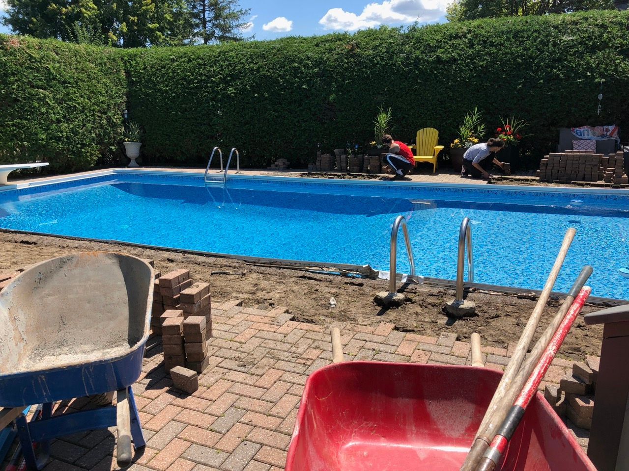 Construction d'une piscine : des ouvriers posent des briques autour d'une piscine bleue. Ciel bleu, haie verte.