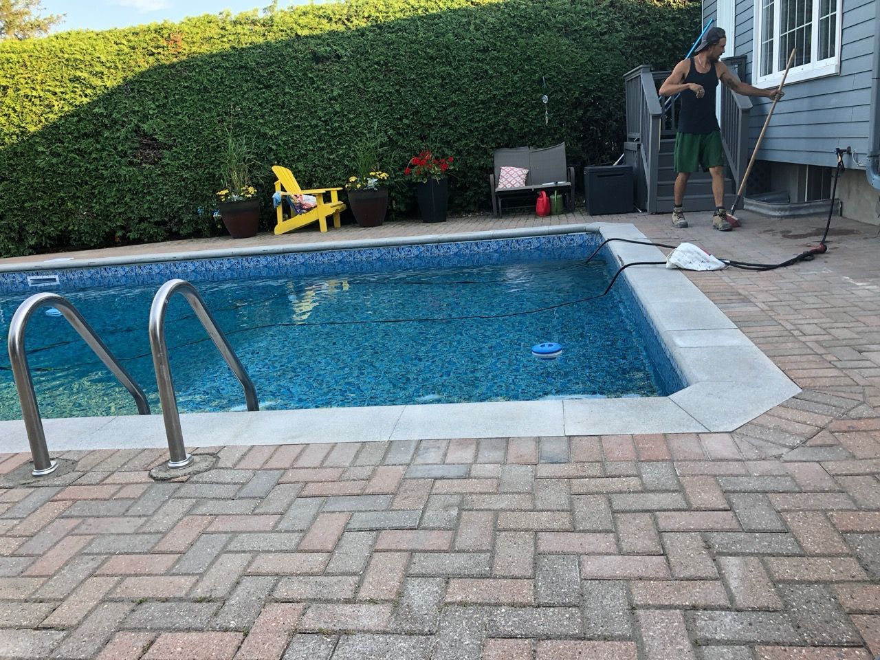 Personne nettoyant une piscine carrelée bleue dans une cour arrière avec patio en briques, entourée de verdure.