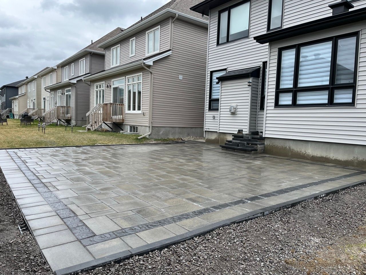 Un patio en pavés gris est bordé de gravier et donne sur des maisons à deux étages avec un revêtement clair.