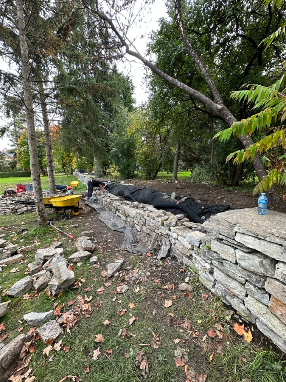 Mur de pierre en construction à côté d'une zone herbeuse, d'arbres et d'outils de travail.