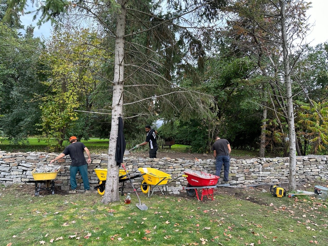 Des hommes construisent un mur de pierre à l'extérieur ; des brouettes jaunes, des outils et des arbres sont visibles.