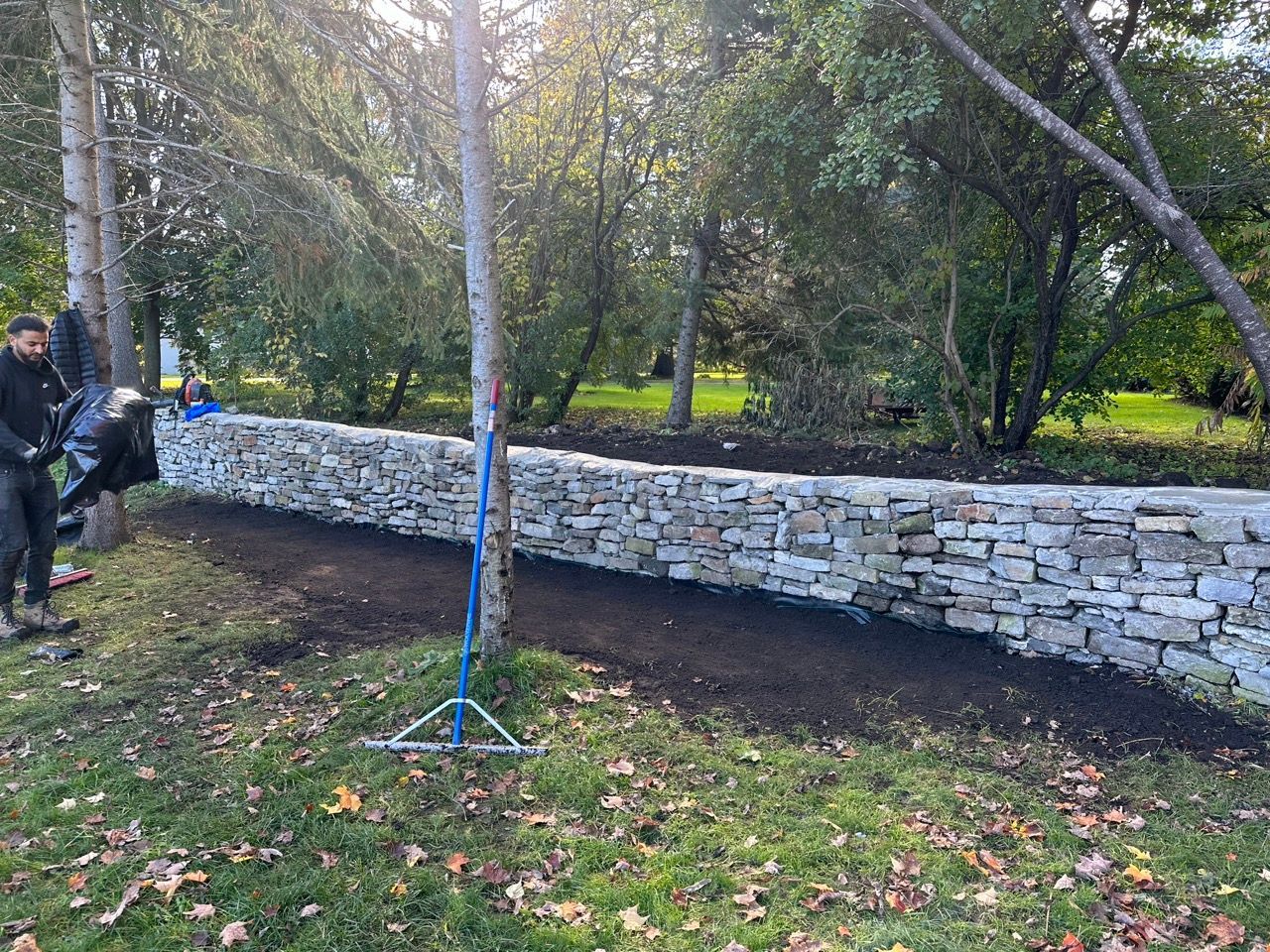 Mur de pierre en construction, avec des ouvriers et des arbres dans un cadre extérieur herbeux.