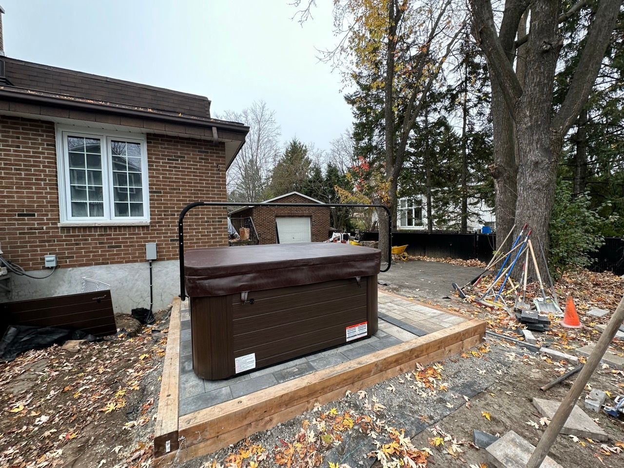 Spa sur une dalle de béton, à côté d'une maison en briques. Couverture et balustrade marron. Extérieur, journée nuageuse.