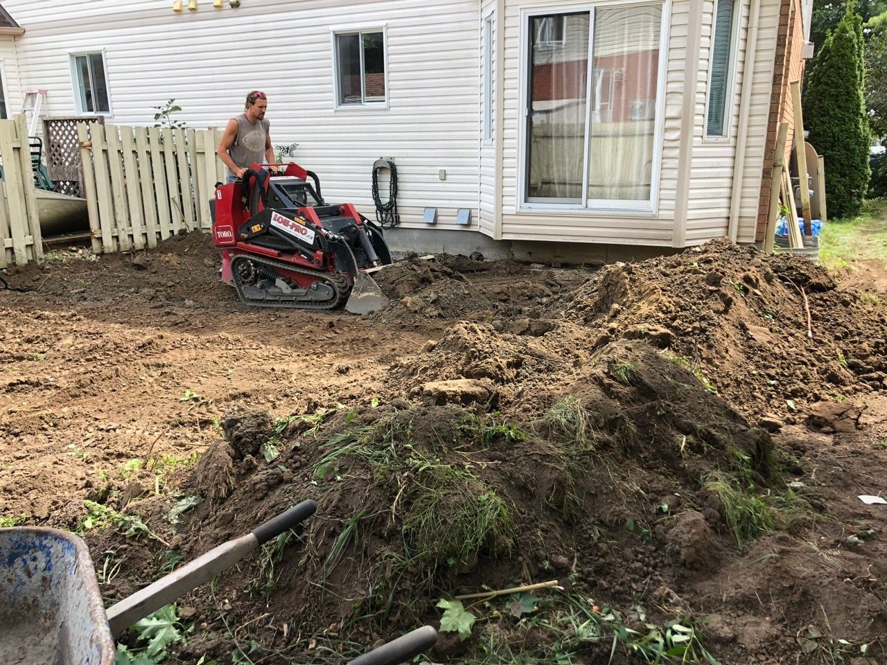 Un homme utilise une mini-pelle pour creuser un terrain près d'une maison. Des tas de terre au premier plan.