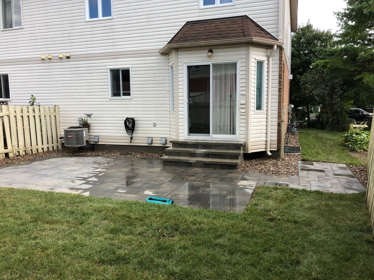 Cour arrière avec patio, marches menant à une porte coulissante en verre, herbe verte et revêtement blanc.
