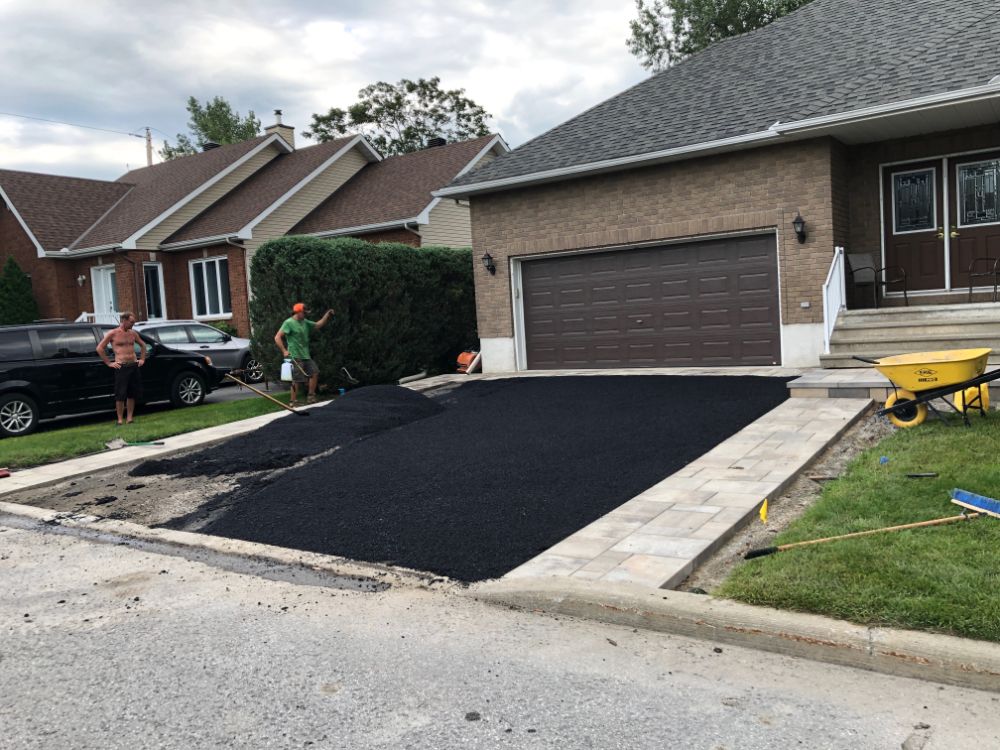 Pavage de l'allée en cours ; deux ouvriers sur le chantier. Un nouvel asphalte noir a été posé à côté d'une bordure en béton clair.