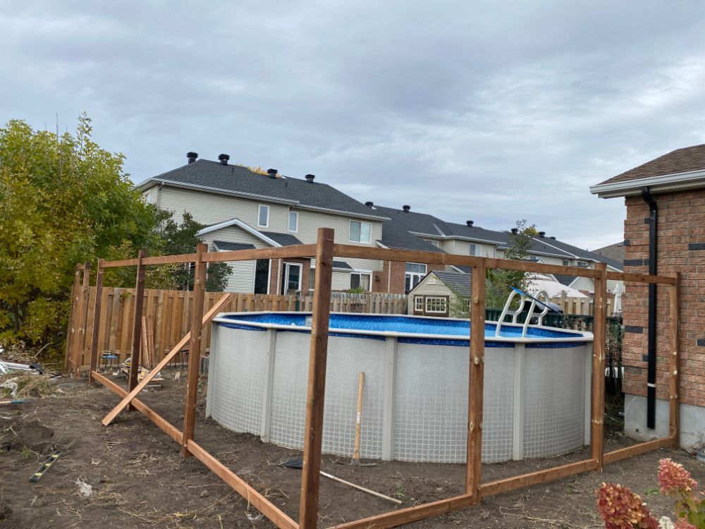 Piscine hors sol entourée d'une clôture en bois sous un ciel nuageux. Des maisons sont visibles en arrière-plan.