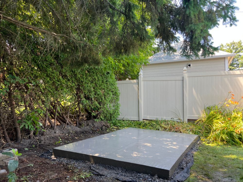 Une dalle de béton grise, rectangulaire et plate, dans un jardin. Feuillage vert et clôture blanche en arrière-plan.