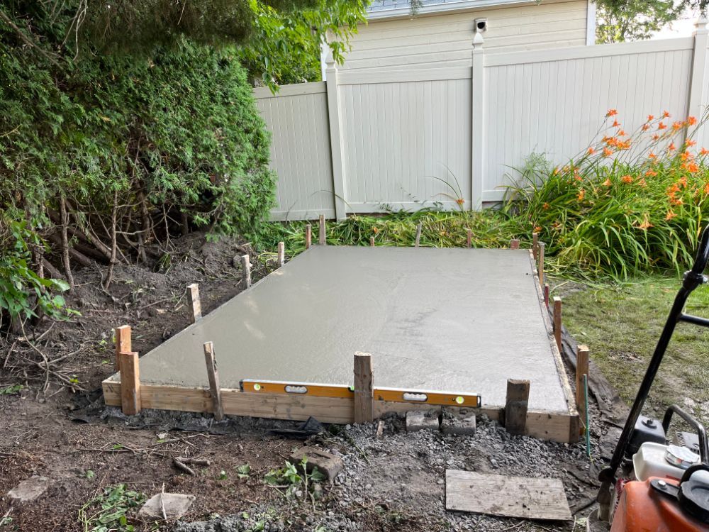 Dalle de béton fraîchement coulée dans des coffrages en bois, dans un jardin, à côté d'une clôture blanche.