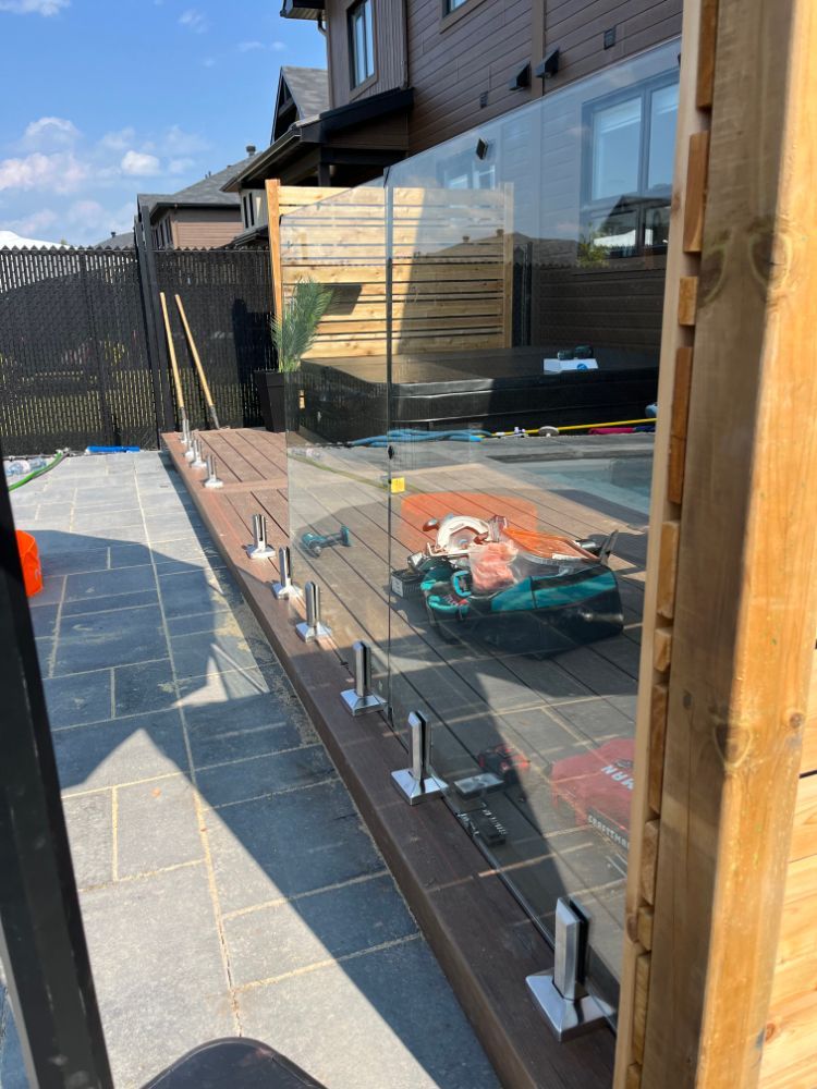 Garde-corps en verre installé sur une terrasse en bois près d'une piscine. Journée ensoleillée, jardin.