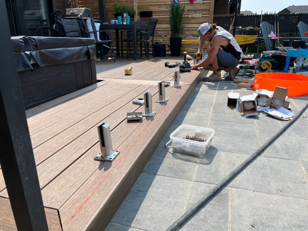 Un homme installe des poteaux de garde-corps en verre sur une terrasse en bois. Cadre extérieur ensoleillé.