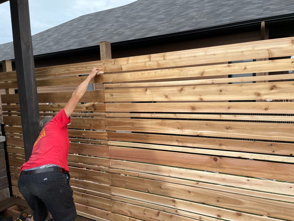 Personne construisant une clôture horizontale en bois à l'extérieur, portant une chemise rouge et un pantalon noir.
