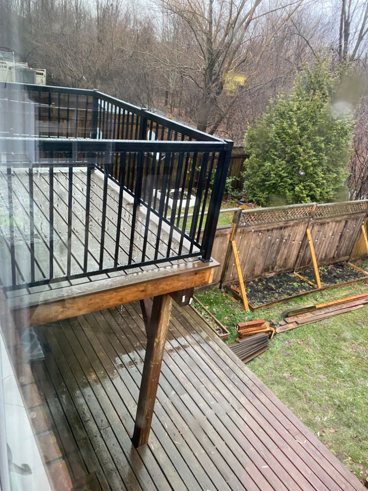 Terrasse en bois avec garde-corps noir, donnant sur cour arrière avec clôture et verdure.