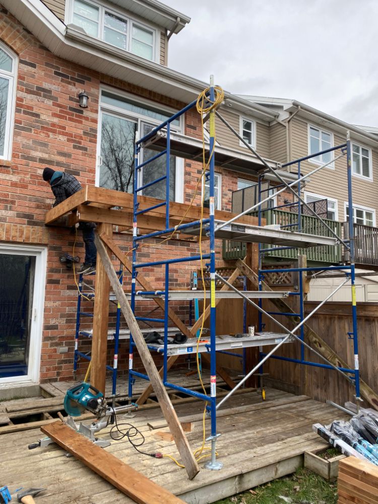 Un homme sur un échafaudage travaille sur un pont attaché à une maison en briques, ciel nuageux au-dessus.
