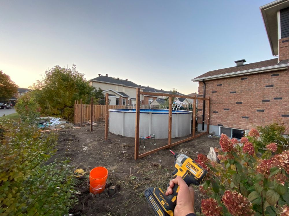 Une personne utilise une perceuse pour construire une clôture autour d’une piscine hors sol dans une cour avec une maison en briques.