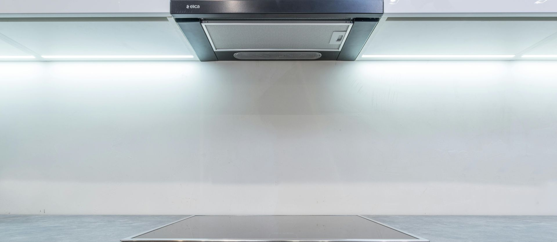A modern kitchen featuring a stainless steel oven and stove top below a range hood and white cabinets with under-glow light.