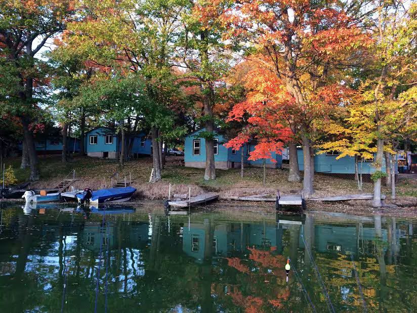 Lakeside cabins with autumn trees reflected in calm water, with small boats and docks along the shore