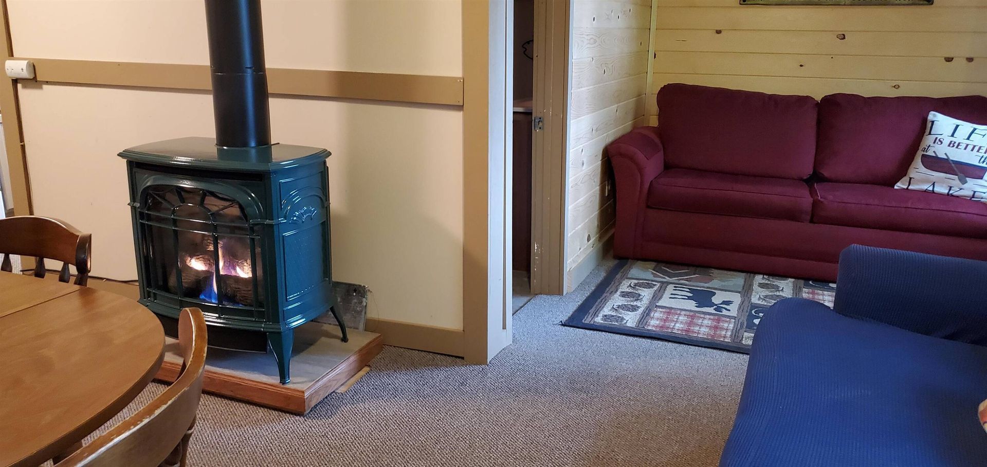 Cozy living room with a blue wood stove, maroon sofa, blue chair, and dining table edge visible