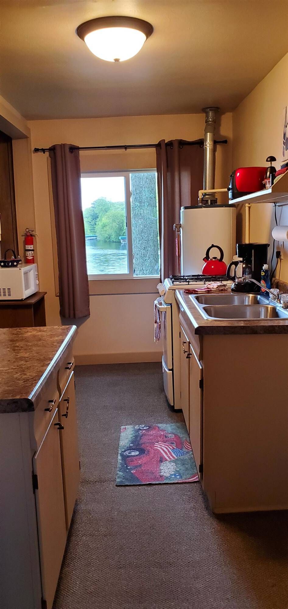 Narrow kitchen with beige cabinets, sink, shelves, and a window at the far end.