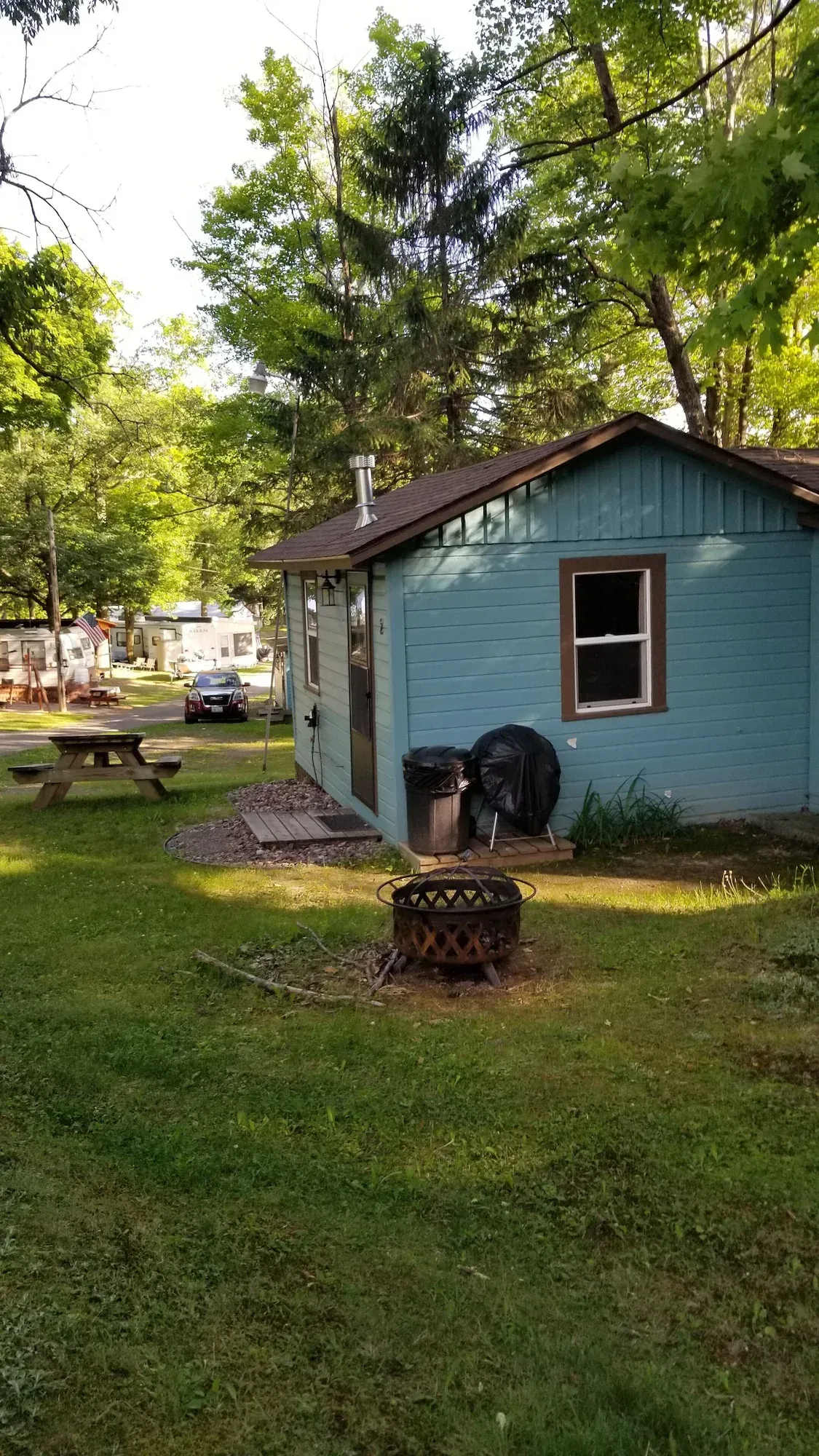 Small blue shed in a wooded yard with picnic tables and grass in the foreground