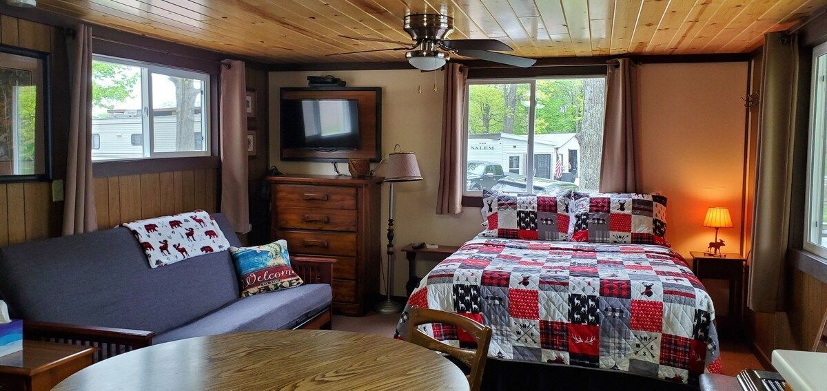 Cozy cabin bedroom with patchwork quilt bed, sofa, TV, and large windows overlooking trees