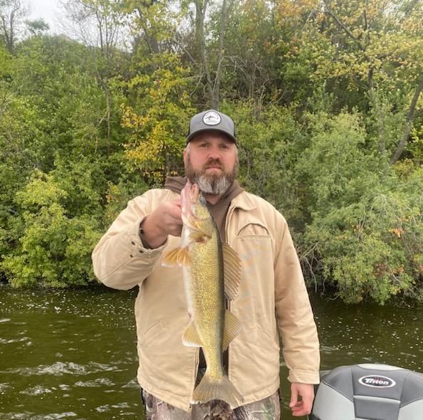 Man holding a large fish by a river, wearing a tan jacket and black cap, with green trees behind him