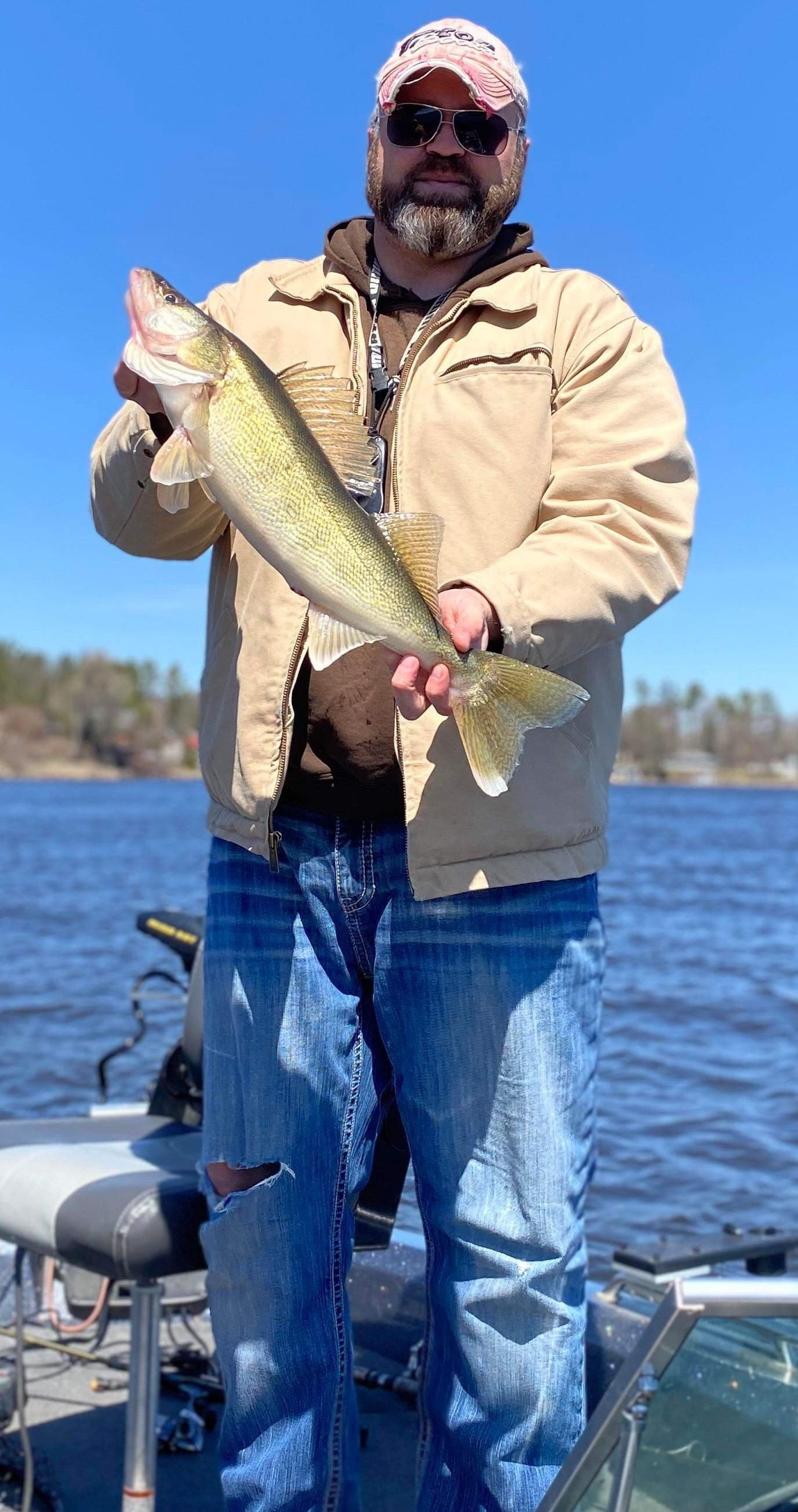 Person holding a large fish on a boat on a lake, wearing a tan jacket, jeans, and a red cap.