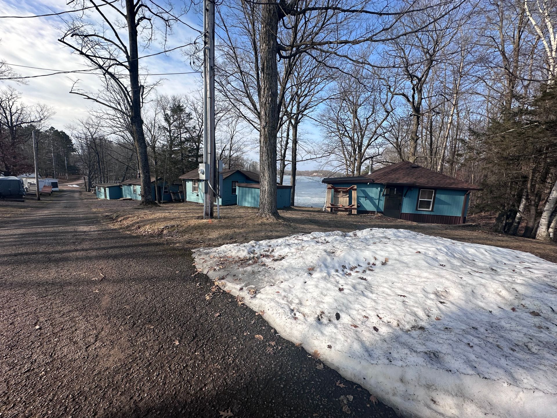 Snow-covered ground beside a gravel path, leafless trees, and a small blue building near a lakeshore.