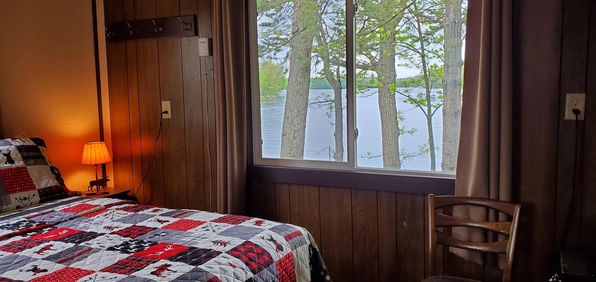 Cozy bedroom with red patterned quilt, wooden walls, lamp, and large window overlooking trees and water