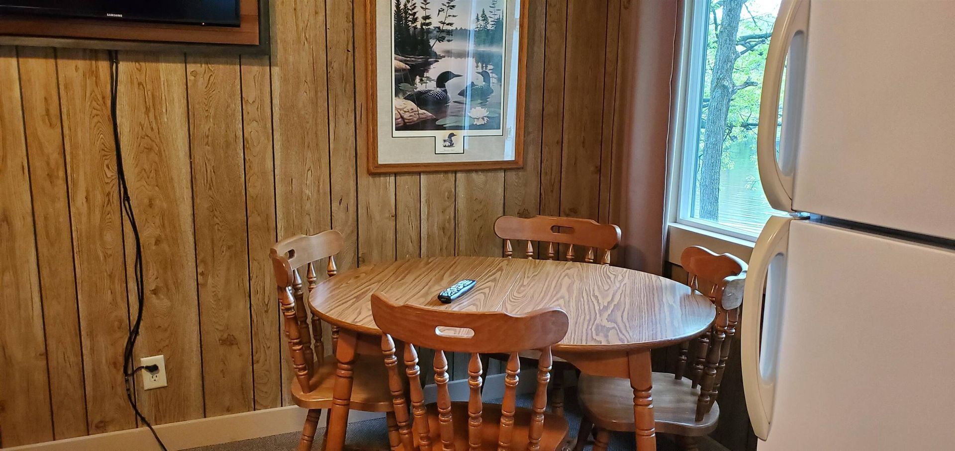 Small wooden dining nook with round table, chairs, wall art, and a refrigerator by the window.