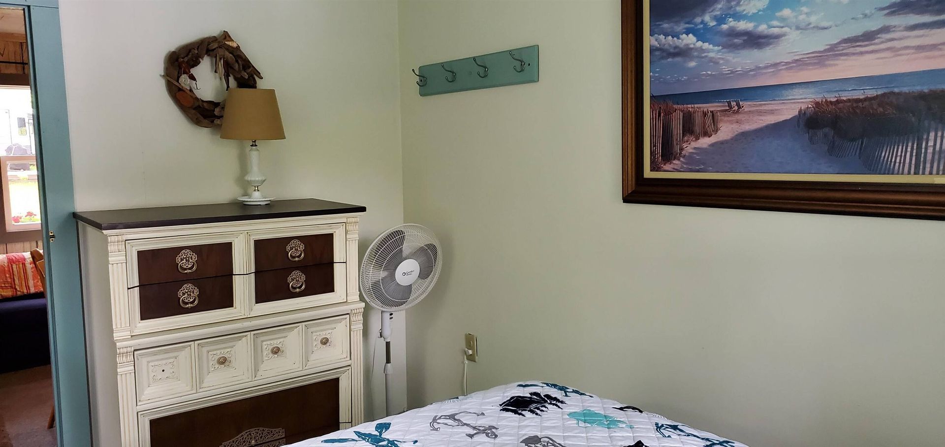 Cozy bedroom corner with a lamp, fan, dresser, framed seaside painting, and patterned bedspread.