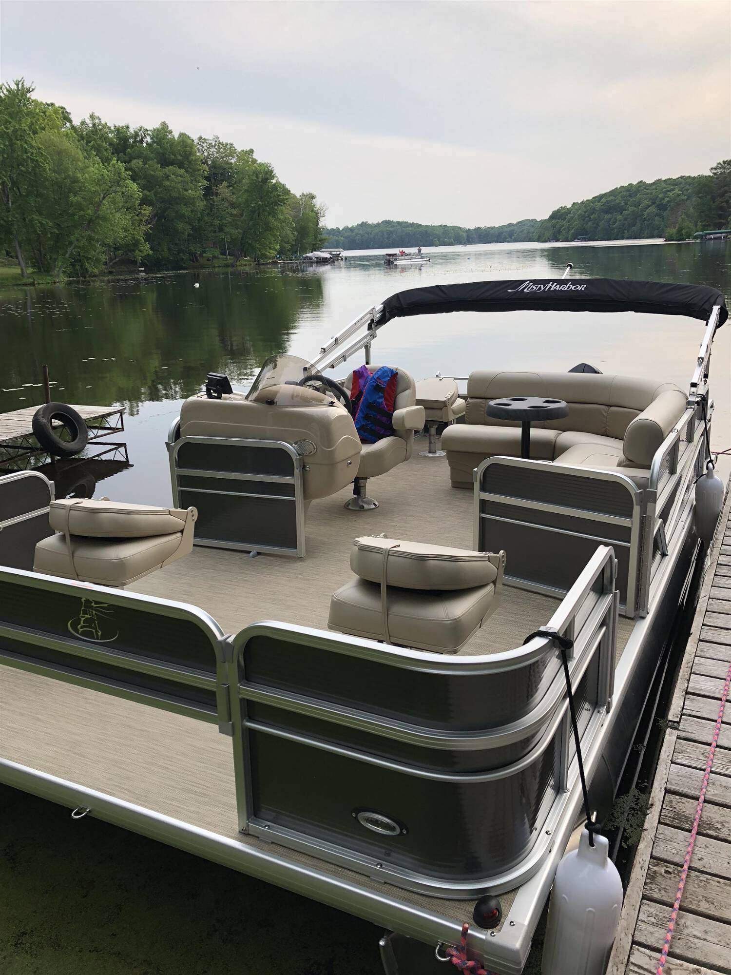 Pontoon boat docked on a calm lake beside a wooded shoreline under an overcast sky