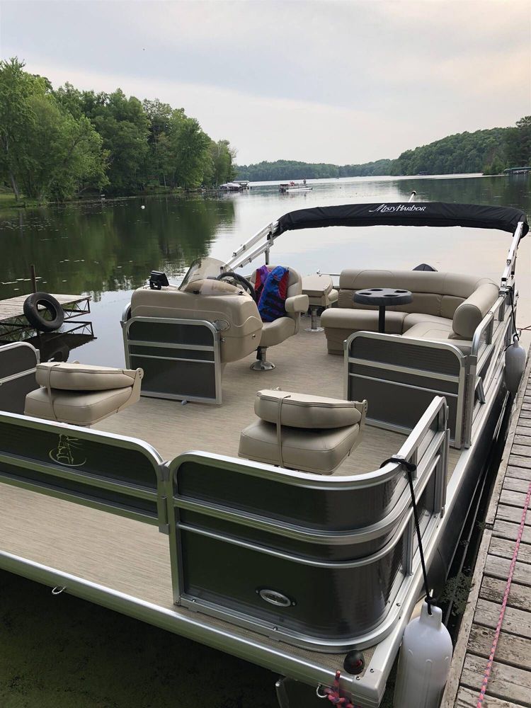Pontoon boat docked on a calm lake, with green forested shoreline and overcast sky.
