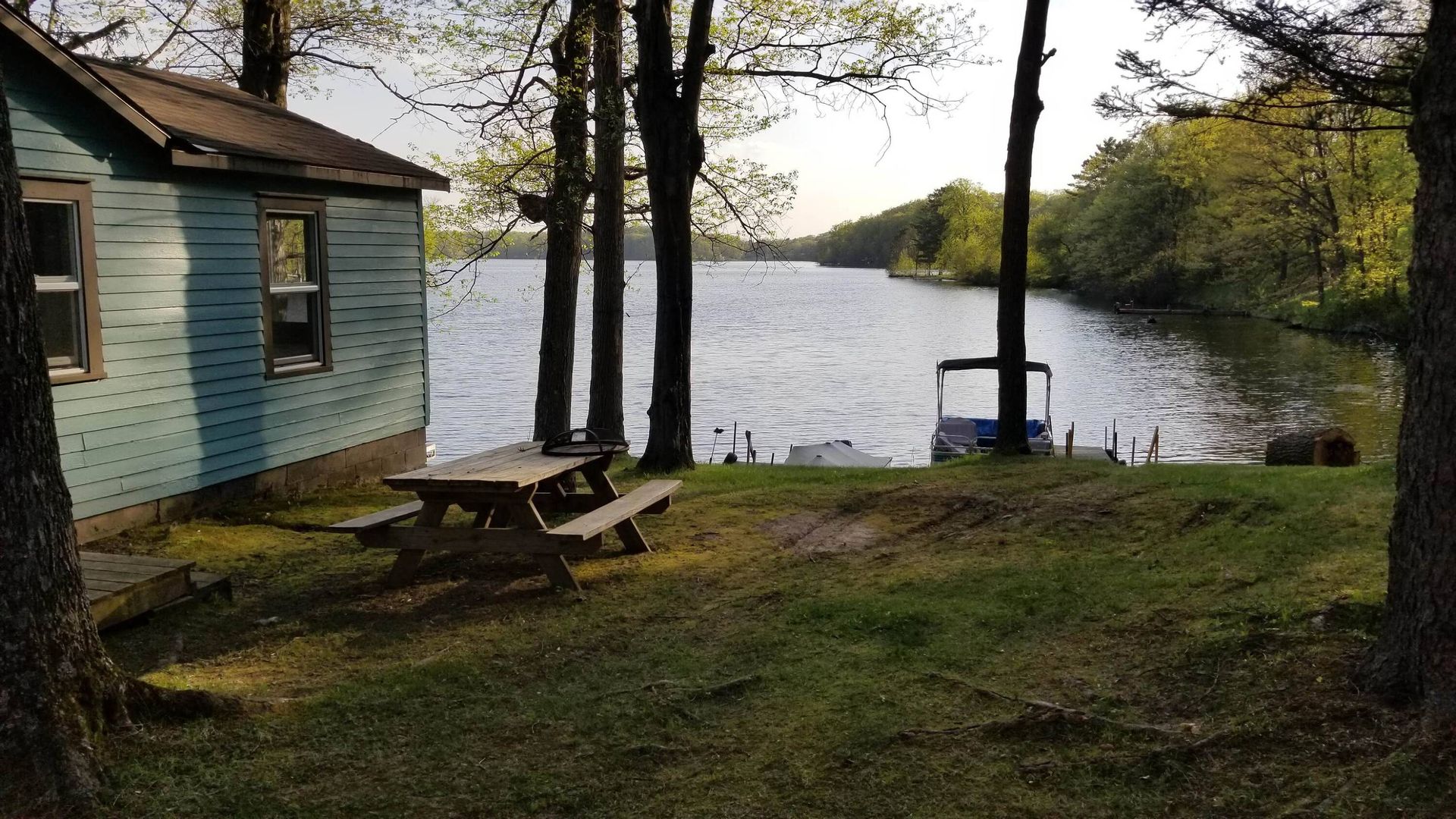 Small blue cabin by a lakeside with picnic tables, trees, and a dock at sunset.