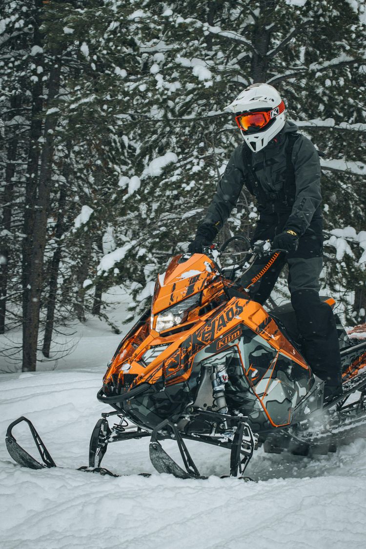 Person riding an orange snowmobile in a snowy forest, wearing black gear and a white helmet with orange goggles