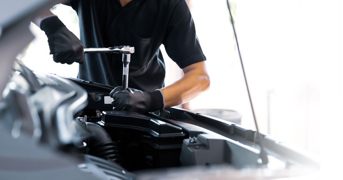 Mechanic in black shirt and gloves, using a wrench on a car engine in a well-lit shop.