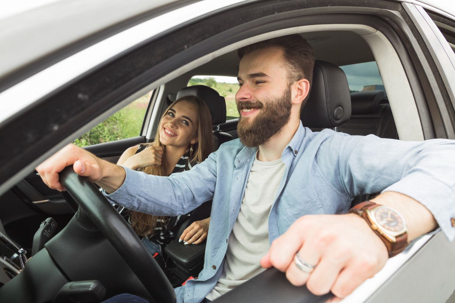 Un hombre conduce un coche, sonriendo, con una mujer en el asiento del copiloto que también sonríe.